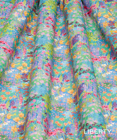 A close-up of draped fabric with a dense, multicolor floral and botanical print on a light blue-green background. The print features pink, yellow, orange, purple, and green flowers and leaves.