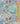 A close-up of a colorful floral fabric print with a gold ruler along the right side for scale. The pattern features a dense, vibrant wildflower meadow with flowers in pink, yellow, orange, purple, and blue on a light blue background.