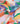 A close-up of draped Liberty Fabrics featuring a colorful geometric pattern with abstract shapes in orange, green, blue, purple, pink, and yellow on a white base.