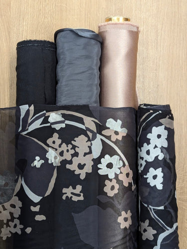 Silk chiffon fabric with a foil printed floral design in champagne, silver and grey on a black base, shown overlaid on three bolts of fabric in solid colours same as the printed florals.