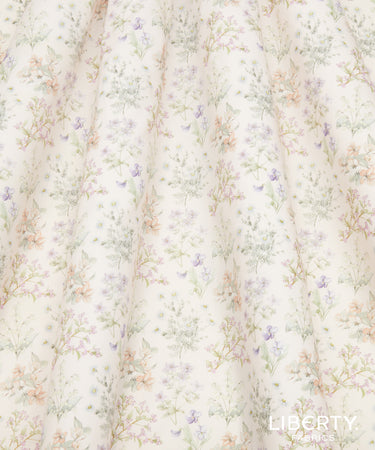 A close-up of a fabric surface with a delicate floral print on a warm off-white background. The pattern features small pastel flowers and leaves in soft purple, peach, and sage green tones, arranged in vertical folds.