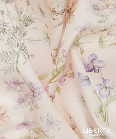 A close-up of Liberty Fabrics floral print fabric in a soft blush pink color, featuring delicate watercolor-style purple flowers, green leaves, and peach blossoms, with the 'LIBERTY FABRICS' logo in the bottom right corner.