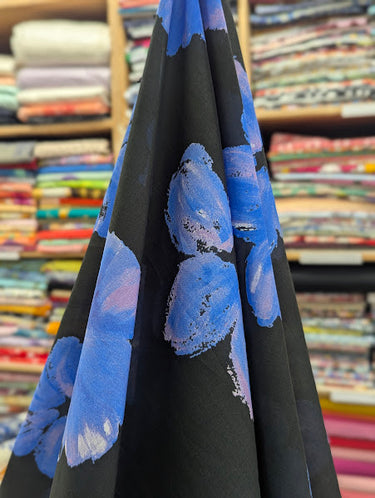 Printed pure cotton fabric, with floral patterns in blue and lilac on a black base, shown draped within a shop setting.