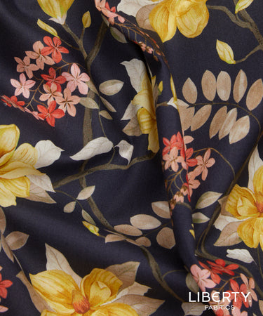 A close-up of a draped fabric with a dark navy background and a floral pattern featuring yellow, pink, and red blossoms with beige leaves. The brand name 'LIBERTY FABRICS' is visible in the bottom right corner.