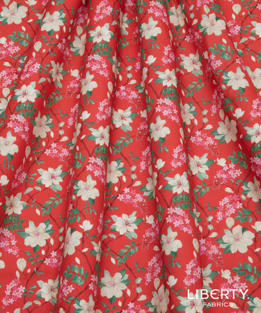 A close-up of a red fabric with a floral print featuring white and pink blossoms and green leaves, with a visible Liberty Fabrics logo in the bottom right corner.