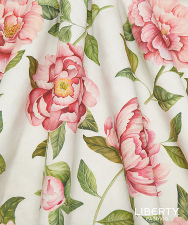 A close-up of a floral fabric print featuring large pink peony flowers and green leaves on a warm off-white background, with the Liberty Fabrics logo in the bottom right corner.