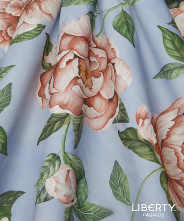 A close-up of a light blue fabric with a large-scale floral print featuring peach-pink peonies and green leaves. The brand name 'LIBERTY.