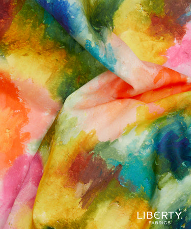 A close-up of colorful tie-dye or watercolor-patterned fabric with blended shades of yellow, pink, blue, green, orange, and white. The Liberty Fabrics logo appears in the bottom right corner.