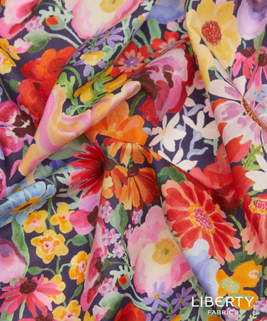 A close-up of draped fabric featuring a vibrant watercolor floral print on a dark navy background. The print includes large flowers in pink, red, orange, yellow, and white, interspersed with green leaves and purple accents.