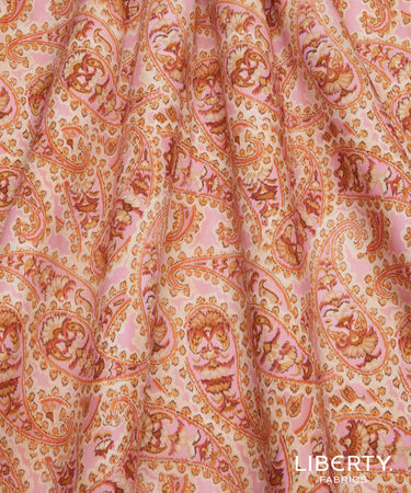 A close-up of a draped fabric with a classic paisley pattern in soft pink, peach, and orange tones, with a 'LIBERTY FABRICS' logo in the bottom right corner.