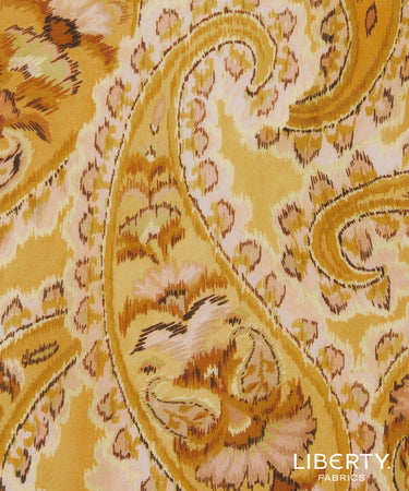 A close-up of a Liberty Fabrics fabric pattern featuring a warm yellow and beige paisley design with brown and pink accents.