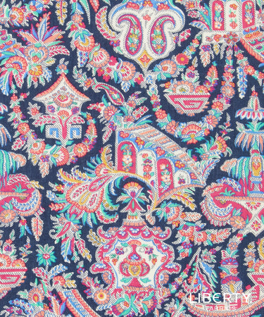 A close-up of a colorful paisley and floral patterned fabric with a dark navy background and bright multicolor motifs including pink, orange, teal, yellow, and white. A small 'Liberty Fabrics' logo watermark is visible in the bottom right corner.