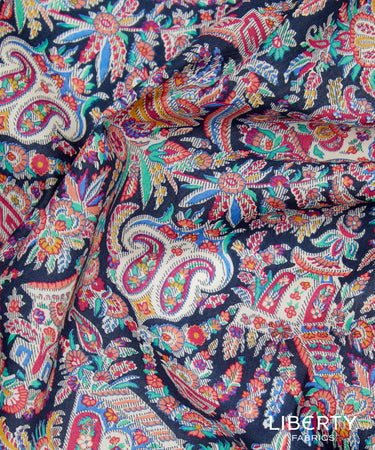 A close-up of a multicolor paisley fabric with intricate floral and ornamental patterns on a dark navy background. The fabric is draped to show its texture and pattern density.