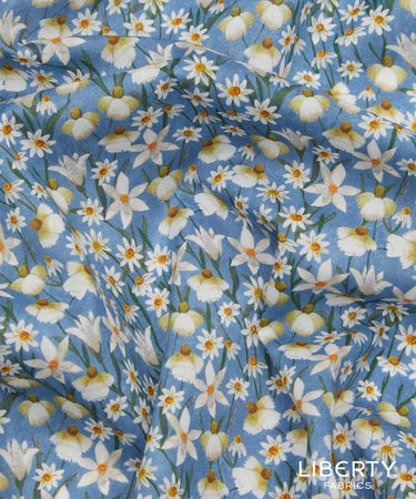 Light blue fabric with an all-over daisy print, featuring white petals, yellow centers, and green stems. A watermark for 'LIBERTY FABRICS' is visible in the bottom right corner.
