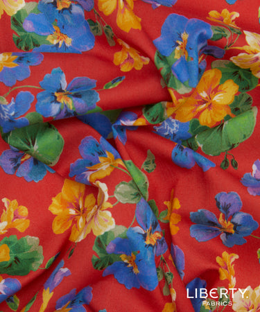 A close-up of a red fabric with a vibrant floral print featuring blue, yellow, and orange flowers with green leaves. The fabric is draped to show its texture and pattern, with a 'LIBERTY FABRICS' logo in the bottom right corner.