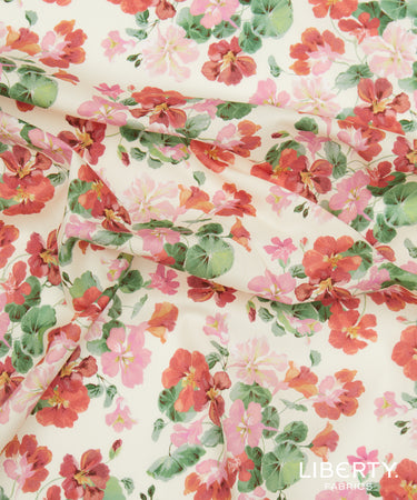 A close-up of draped fabric featuring a watercolor-style floral print with red, pink, and coral flowers and green leaves on a warm cream background. A 'LIBERTY FABRICS' watermark is visible in the bottom right corner.