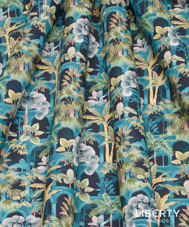 A close-up of draped fabric with a dense tropical print featuring teal, navy, green, beige, and white floral and palm motifs. The Liberty Fabrics logo is visible in the bottom right corner.