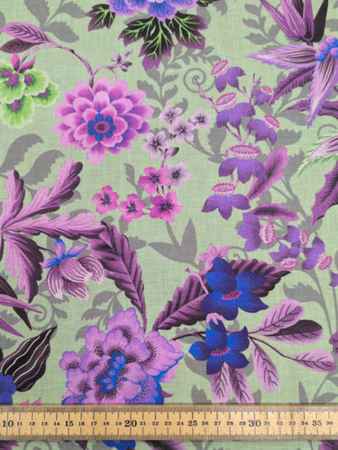 A linen fabric with a bold floral print featuring primarily blue, pink and grey colours on a green background, with a ruler at the bottom for scale.