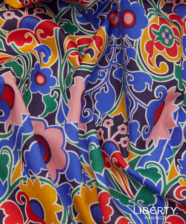 A close-up of a colorful printed fabric with a bold floral and paisley pattern in blue, pink, red, yellow, green, and white, with a visible Liberty Fabrics logo in the bottom right corner.