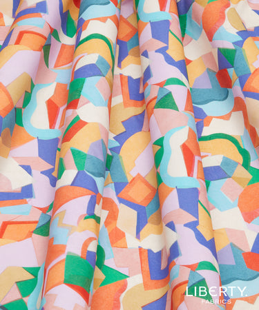 A close-up of draped fabric with a colorful abstract geometric pattern featuring orange, blue, green, yellow, and pink shapes on a white background. 