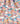 A close-up of draped fabric with a colorful scenic print featuring mountains, clouds, trees, flowers, and stylized figures in pink, blue, white, yellow, and orange tones.