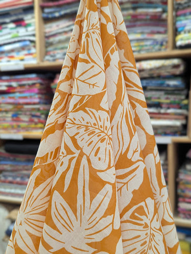 A printed pure cotton fabric with ivory botanical print on a mustard base, shown draped within a shop setting.