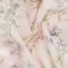 A close-up of Liberty Fabrics floral print fabric in a soft blush pink color, featuring delicate watercolor-style purple flowers, green leaves, and peach blossoms, with the 'LIBERTY FABRICS' logo in the bottom right corner.
