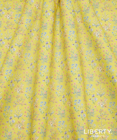 Yellow fabric with a dense, all-over floral and botanical print in shades of blue, pink, and green, featuring a visible 'LIBERTY FABRICS' logo in the bottom right corner.