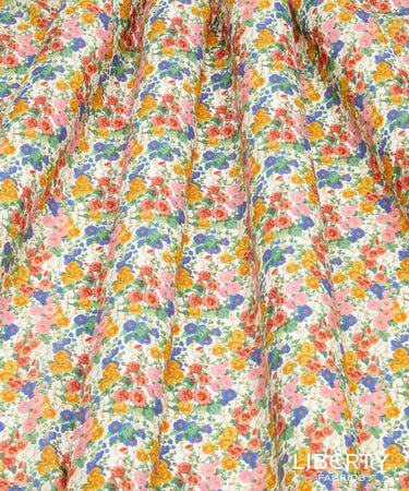 A close-up of a lightweight fabric with a dense, multicolor floral print featuring pink, orange, blue, and yellow flowers on a white background, draped in soft vertical folds. A 'Liberty Fabrics' logo is visible in the bottom right corner.