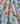A close-up of a light blue fabric with a large-scale floral print featuring peach-pink peonies and green leaves. The brand name 'LIBERTY.