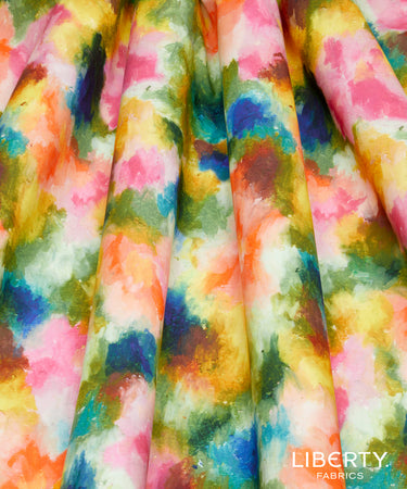 A close-up of draped fabric with a watercolor-style multicolor print featuring pink, yellow, blue, green, orange, and white tones. The Liberty Fabrics logo is visible in the bottom right corner.