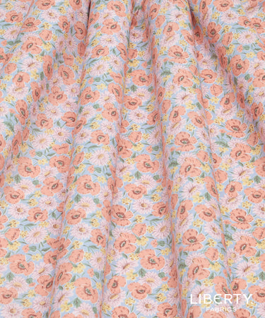 A close-up of a floral fabric with a repeating pattern of peach, pink, yellow, and green flowers on a pale blue background. The fabric is draped with soft vertical folds, and a watermark for 'LIBERTY FABRICS' is visible in the bottom right corner.