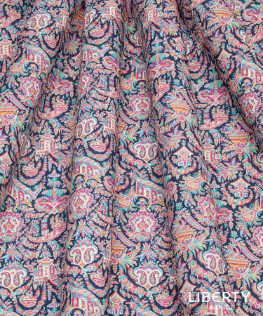 A close-up of draped fabric featuring a detailed, multicolored traditional pattern with pink, orange, teal, and white motifs on a dark navy base. A white 'LIBERTY Fabrics' logo is visible in the bottom right corner.