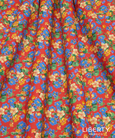 A close-up of a draped fabric with a vibrant red background and an all-over floral print featuring blue, yellow, and green flowers. The Liberty Fabrics logo is visible in the bottom right corner.