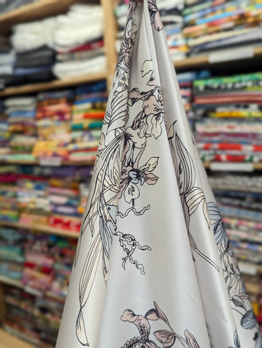 Silvene 100% Silk Satin fabric with a pastel coloured floral pattern and black outlines, shown draped within a shop setting.